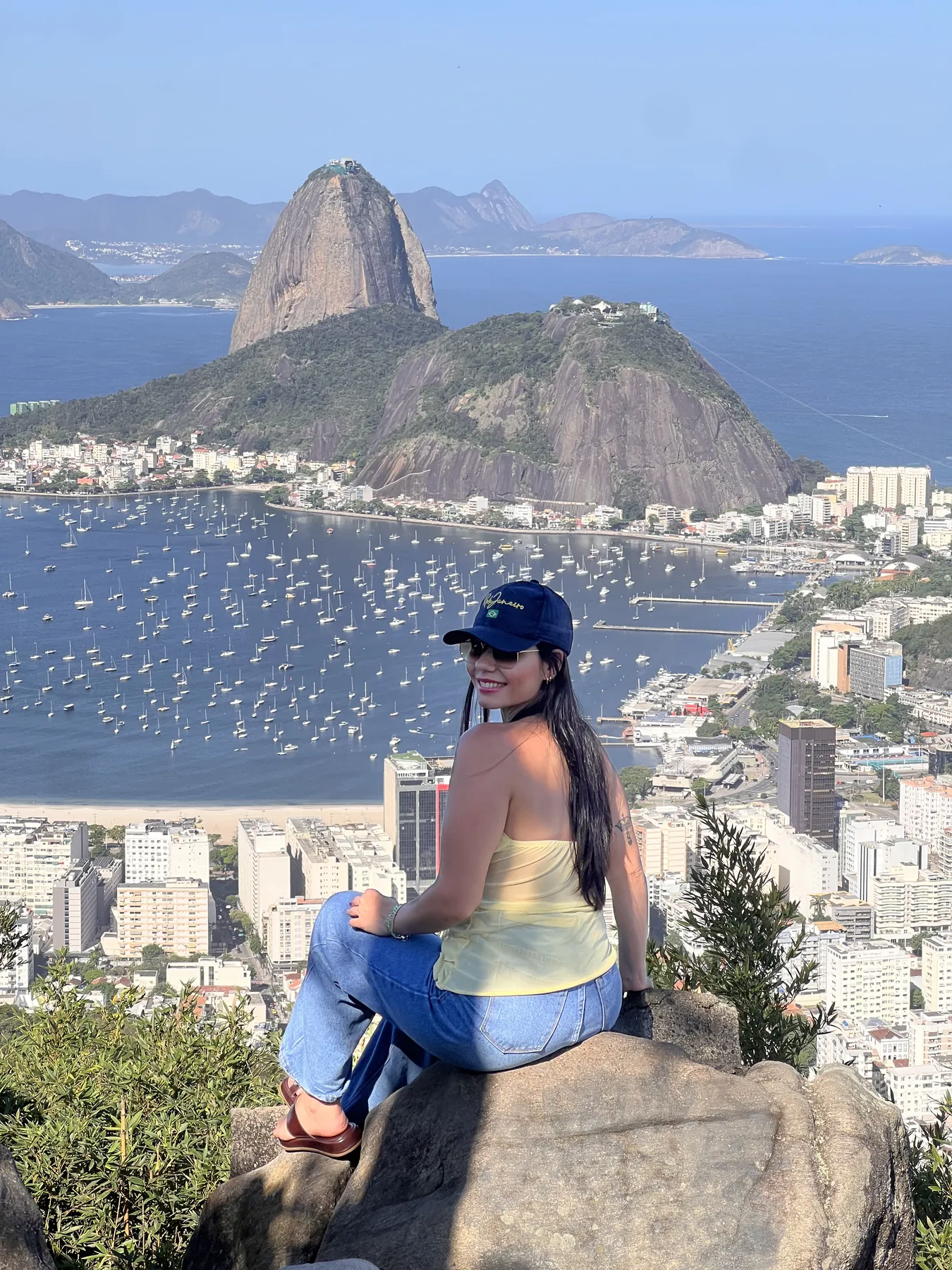 Sabrina sentada em uma pedra de mirante, com a vista do Pão de Açúcar e da Baía de Guanabara ao fundo