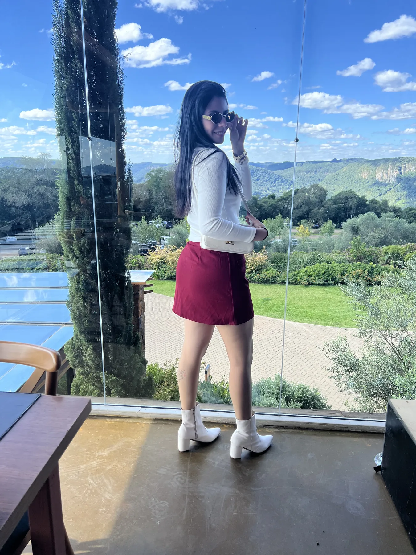 Sabrina de saia vermelha e botas brancas em frente a uma parede de vidro com vista para as montanhas